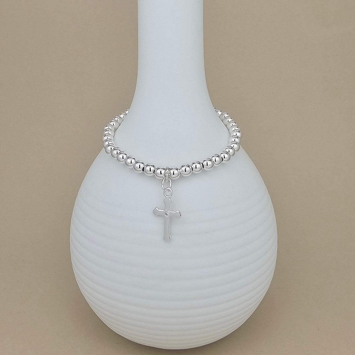 A silver beaded bracelet with a small cross charm, displayed on a white ribbed ceramic vase against a solid beige background.