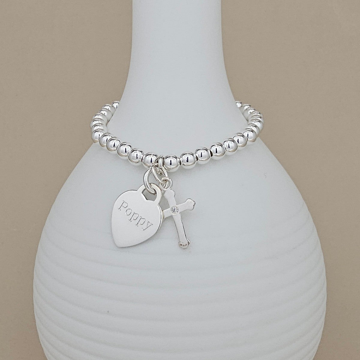 A silver beaded bracelet with a heart-shaped charm engraved with 'Poppy' and a small cross charm, displayed on a white ribbed vase against a beige background.