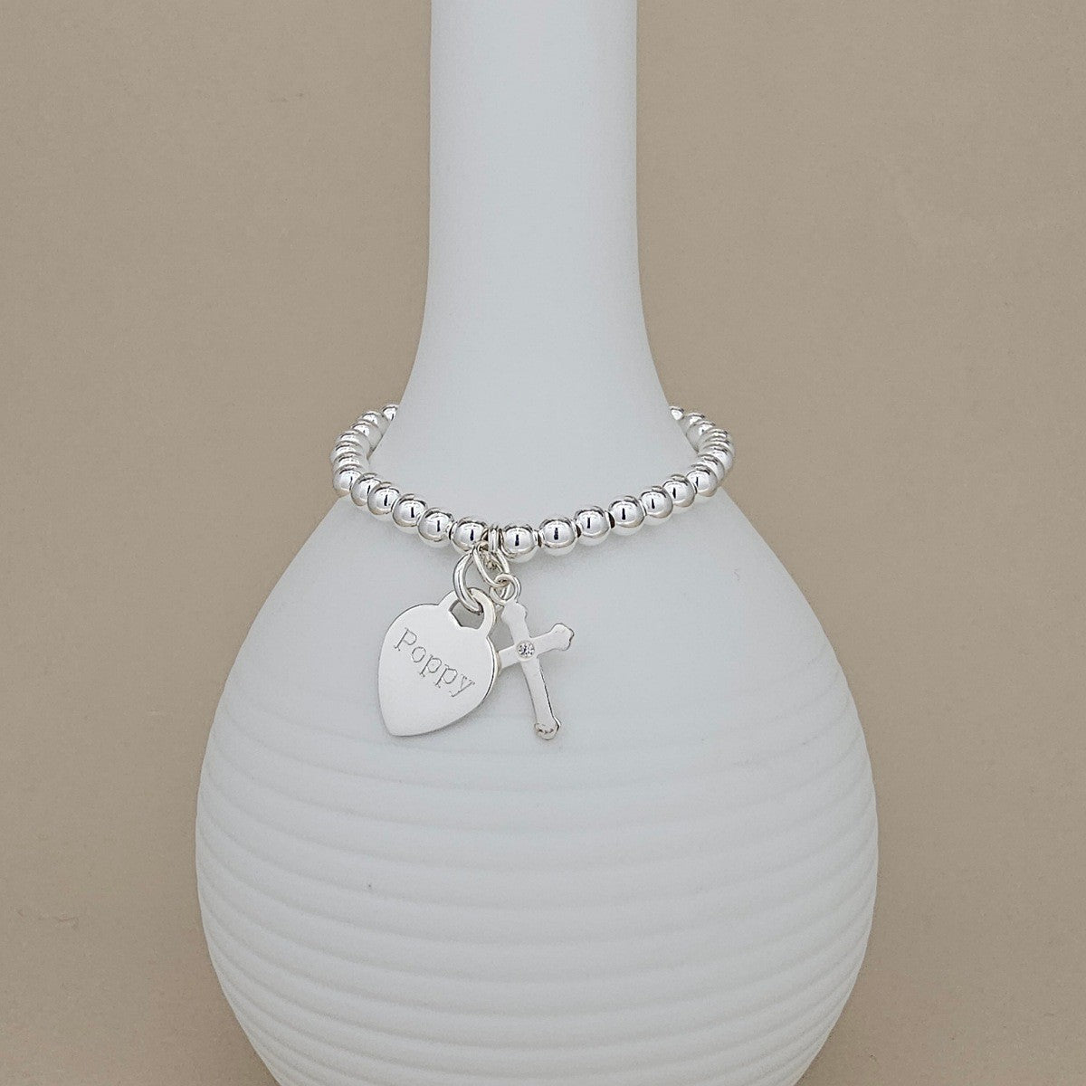 A silver beaded bracelet with a heart-shaped tag engraved with 'Pappy' and a small cross charm, displayed on a white ribbed vase against a beige background.