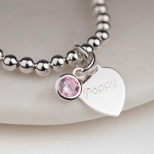 A silver beaded bracelet with a heart-shaped charm engraved with the name 'Poppy' and a round pink gemstone charm, shown on a soft light beige background.