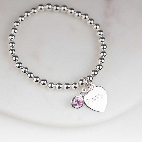 A silver beaded bracelet with a heart-shaped charm engraved with 'poppy' and a small pink gemstone charm, displayed on a white marble surface.