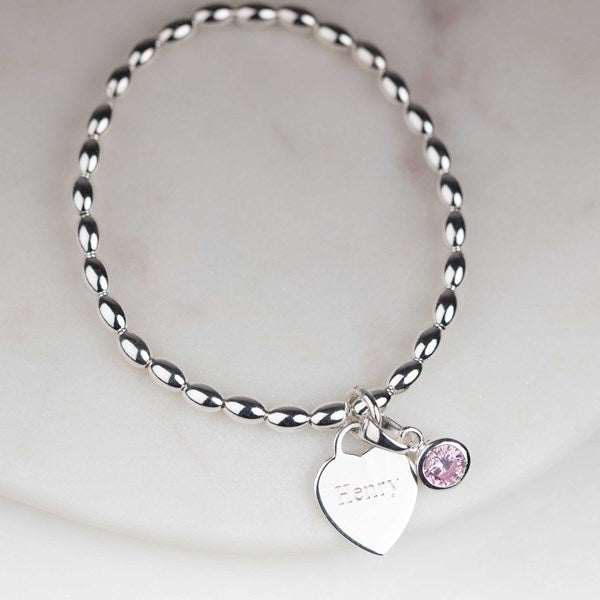 A silver beaded stretch bracelet with a heart-shaped nameplate charm engraved with 'Henry' and a small pink gemstone charm, displayed on a light marble surface.