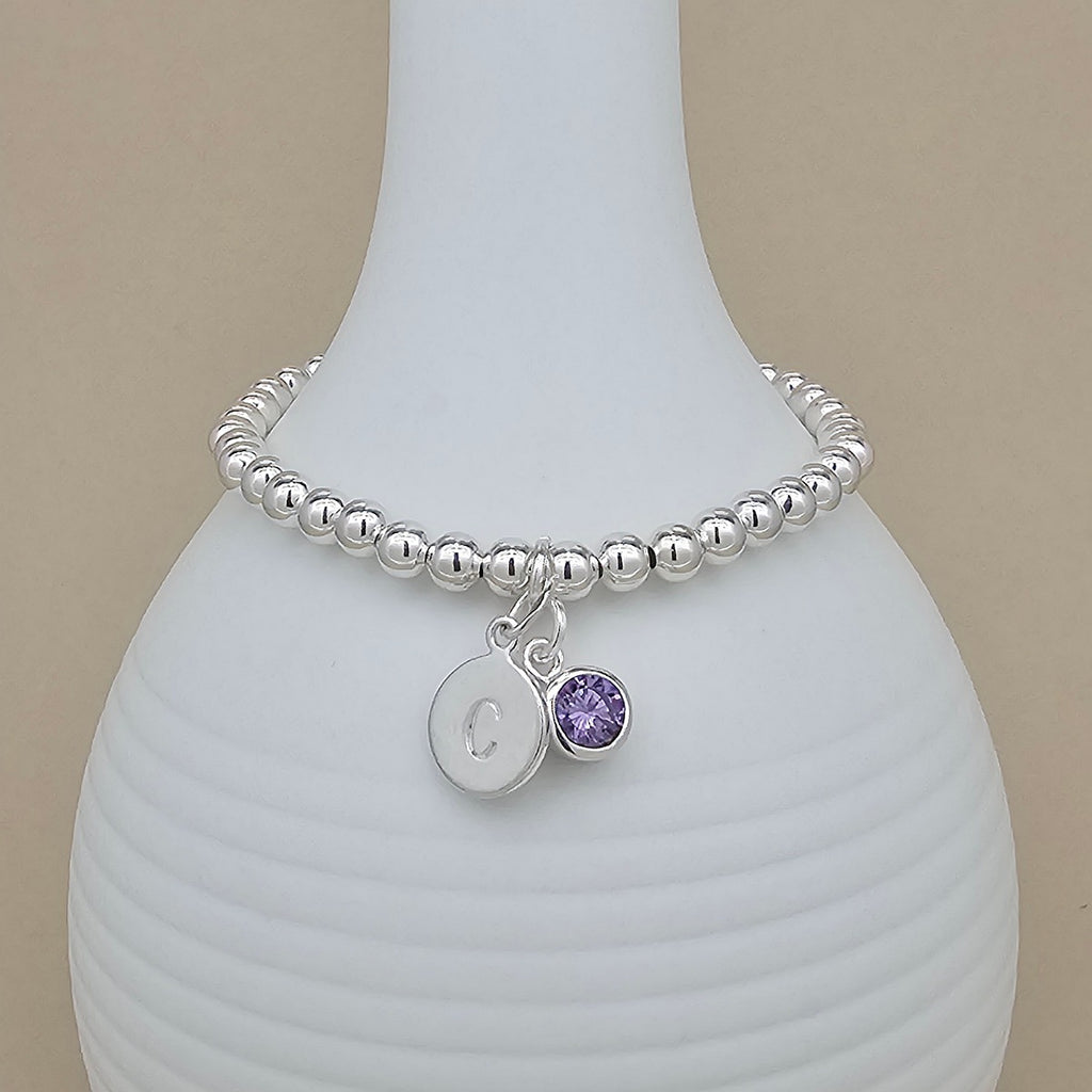 A silver beaded bracelet with a round initial 'C' charm and a small purple gemstone charm, displayed on a white ribbed ceramic vase against a beige background.