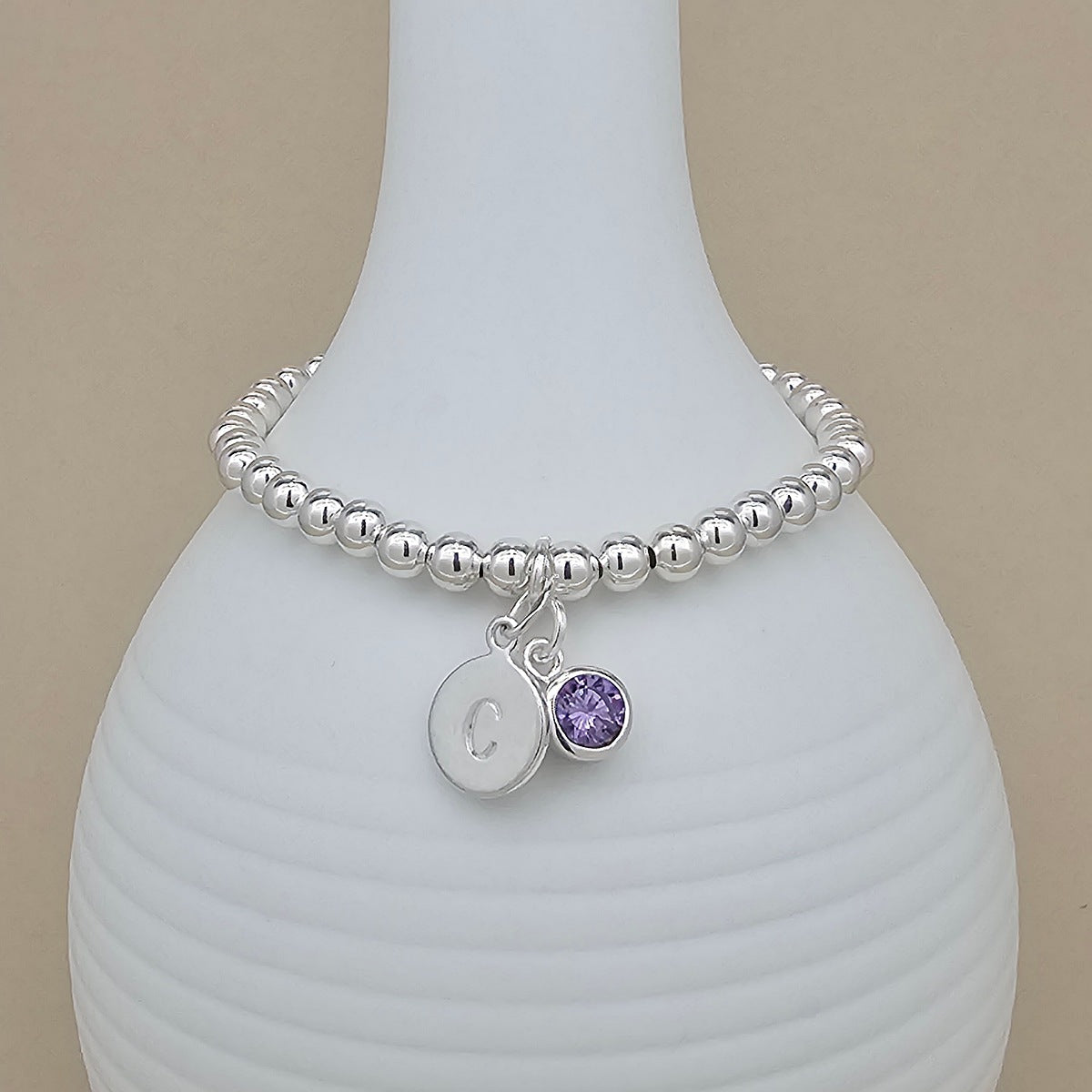 A silver beaded bracelet with a round initial 'C' charm and a small purple gemstone charm, displayed on a white ribbed ceramic vase against a beige background.