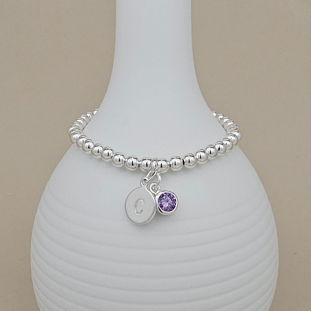 A silver beaded bracelet with a round initial 'C' charm and a small purple crystal charm, displayed on a white ribbed vase against a beige background.