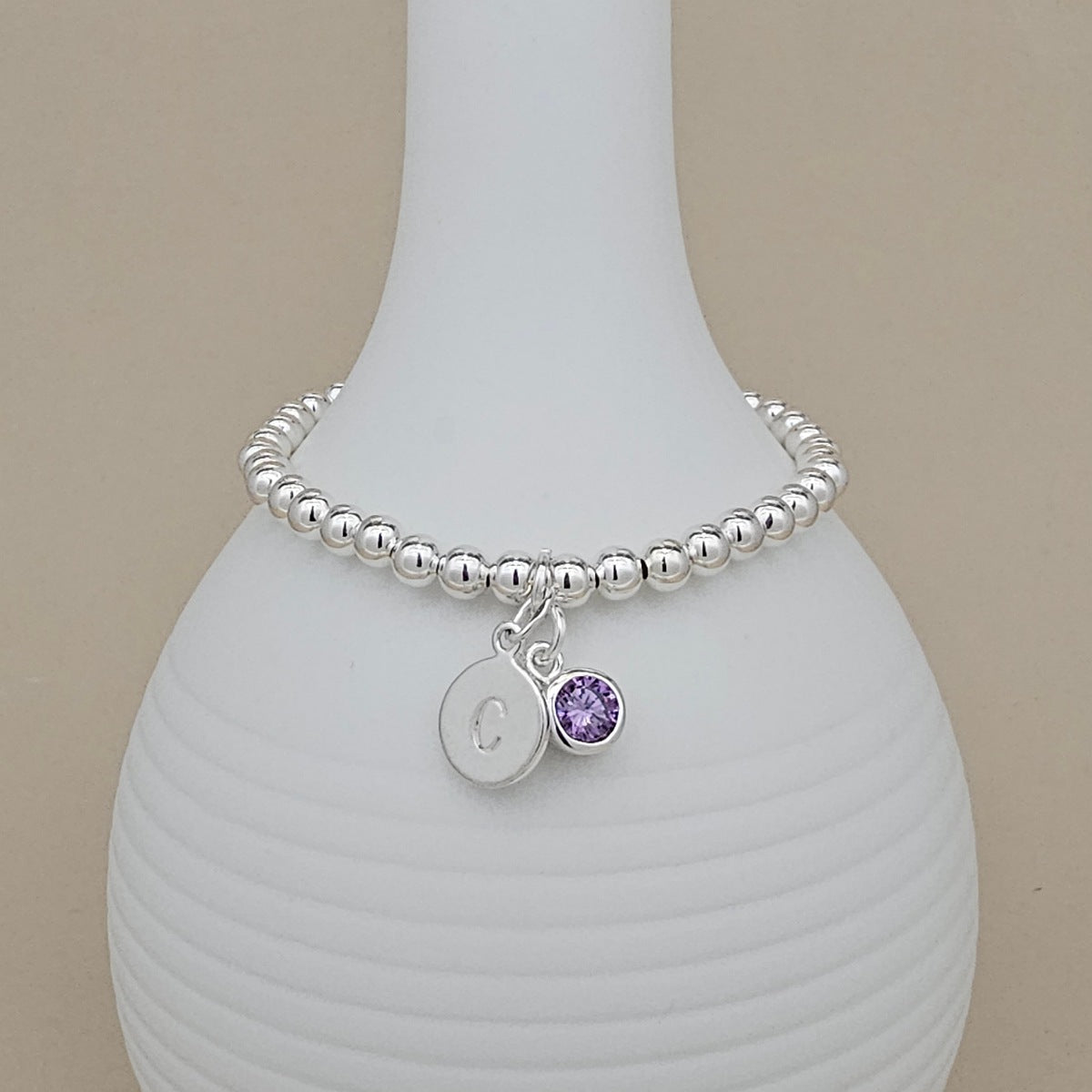 A silver beaded bracelet with a round initial 'C' charm and a small purple crystal charm, displayed on a white ribbed vase against a beige background.