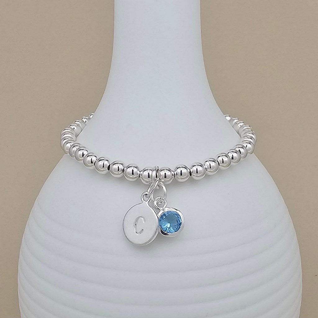 A silver beaded bracelet with a round initial 'C' charm and a blue gemstone charm, displayed on a white ribbed vase against a beige background.