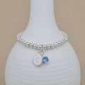 A silver beaded bracelet with a round initial 'C' charm and a blue gemstone charm, displayed on a white ribbed vase against a beige background.