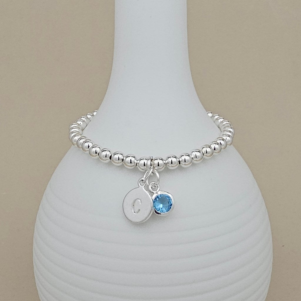 A silver beaded bracelet with a round initial 'C' charm and a light blue gemstone charm, displayed on a white ribbed vase against a beige background.