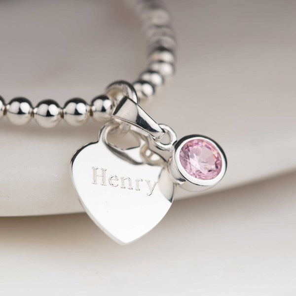 Silver bracelet with a heart-shaped charm engraved 'Henry' and a pink gemstone on a light background.