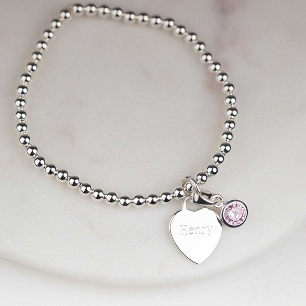 A silver beaded bracelet with a heart-shaped charm engraved with the name 'Henry' and a small round pink crystal accent, displayed on a light marble surface.