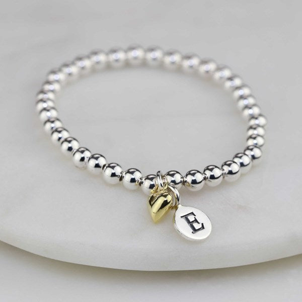 Silver beaded bracelet with gold charm and letter 'E' charm on a white stone surface