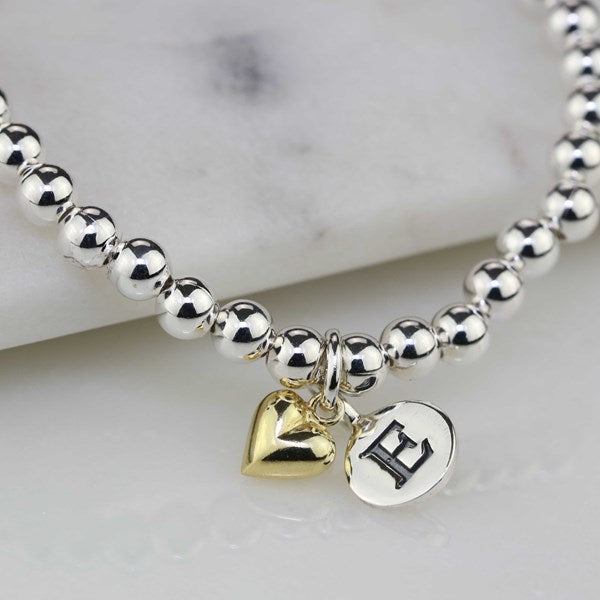 A silver beaded bracelet with two dangling charms: a polished gold heart and a silver oval disc engraved with a black capital letter E, displayed on a white marble surface.