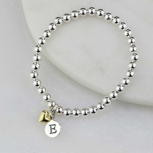 A silver beaded stretch bracelet with a small gold heart charm and a round silver initial 'E' disc charm, displayed on a white marble prop.