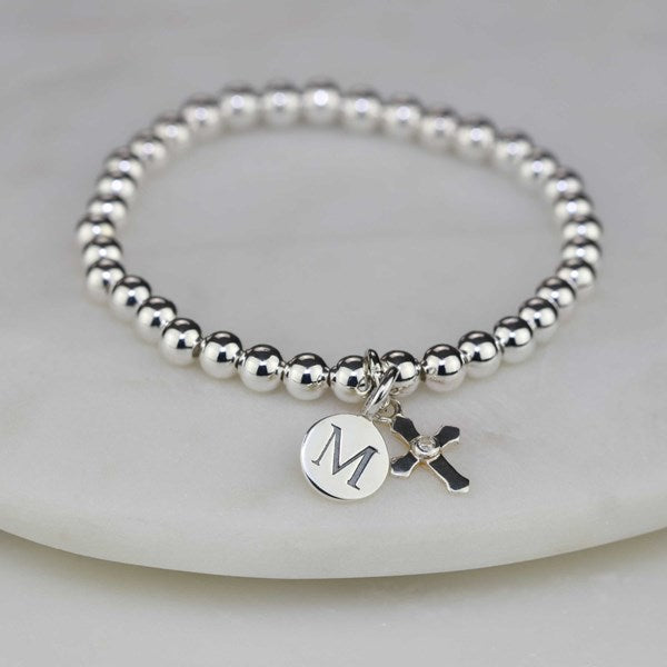 A silver beaded bracelet with a round initial 'M' charm and a small cross charm, displayed on a white stone slab.