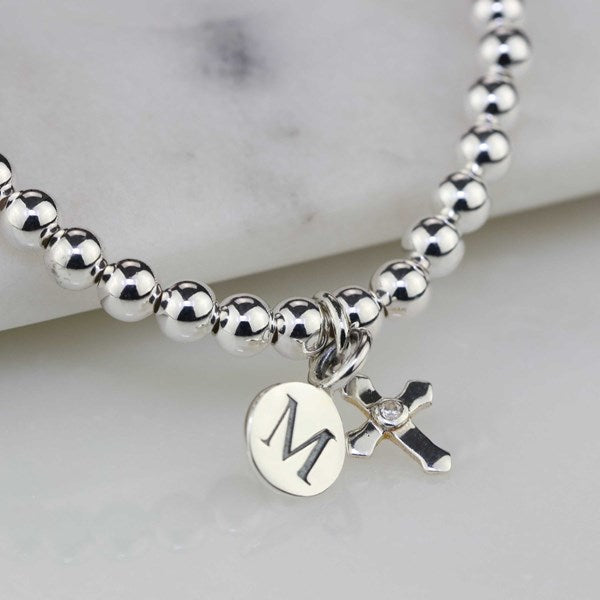 A silver beaded bracelet with a round initial 'M' disc charm and a small cross charm, displayed on a light marble surface.