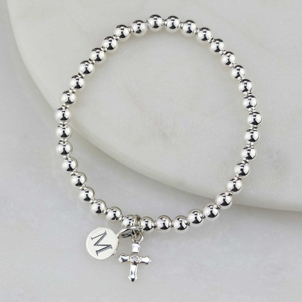 A silver beaded bracelet with a small cross charm and a round engraved initial 'M' disc charm, displayed on a white curved surface.