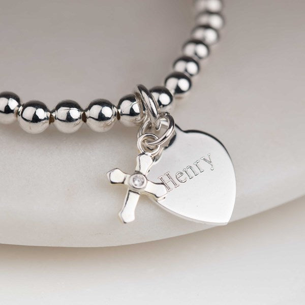 A close-up of a silver beaded bracelet with a heart-shaped charm engraved with the name 'Henry' and a small cross charm with a clear rhinestone, resting on a light beige surface.