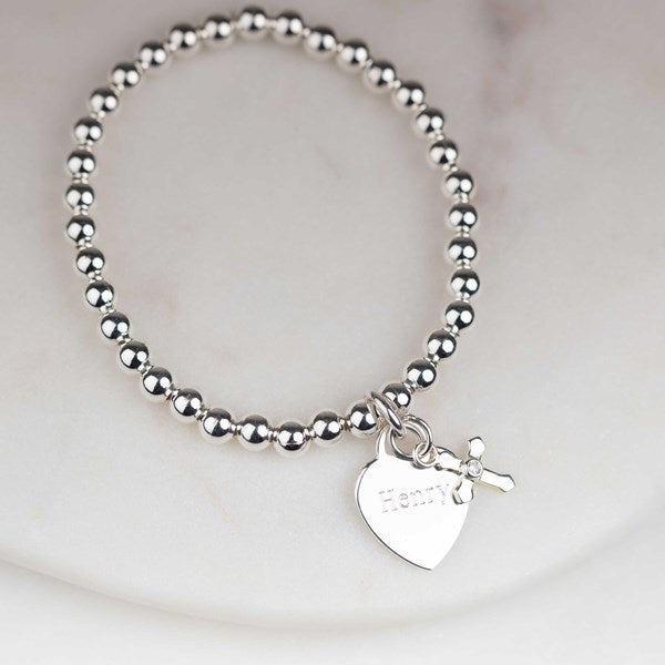 A silver beaded bracelet with a heart-shaped charm engraved with the name 'Henry' and a small cross charm, displayed on a light marble surface.