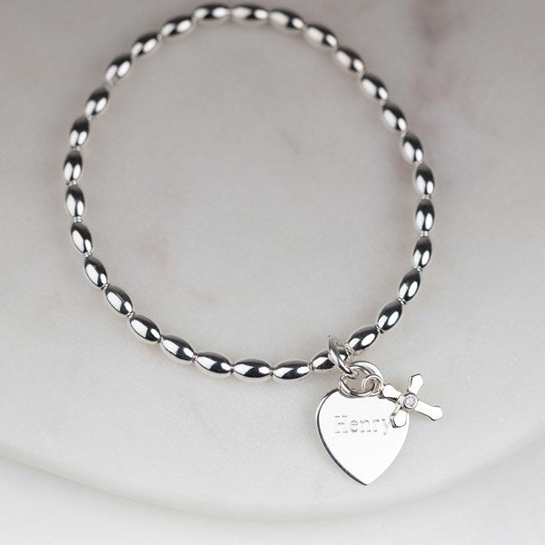A silver bead bracelet with a heart-shaped charm engraved with the name 'Henry' and a small cross charm, displayed on a light marble surface.