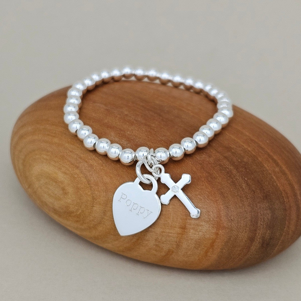 Silver bracelet with heart engraved with the name 'Poppy' and cross charms on a wooden rock
