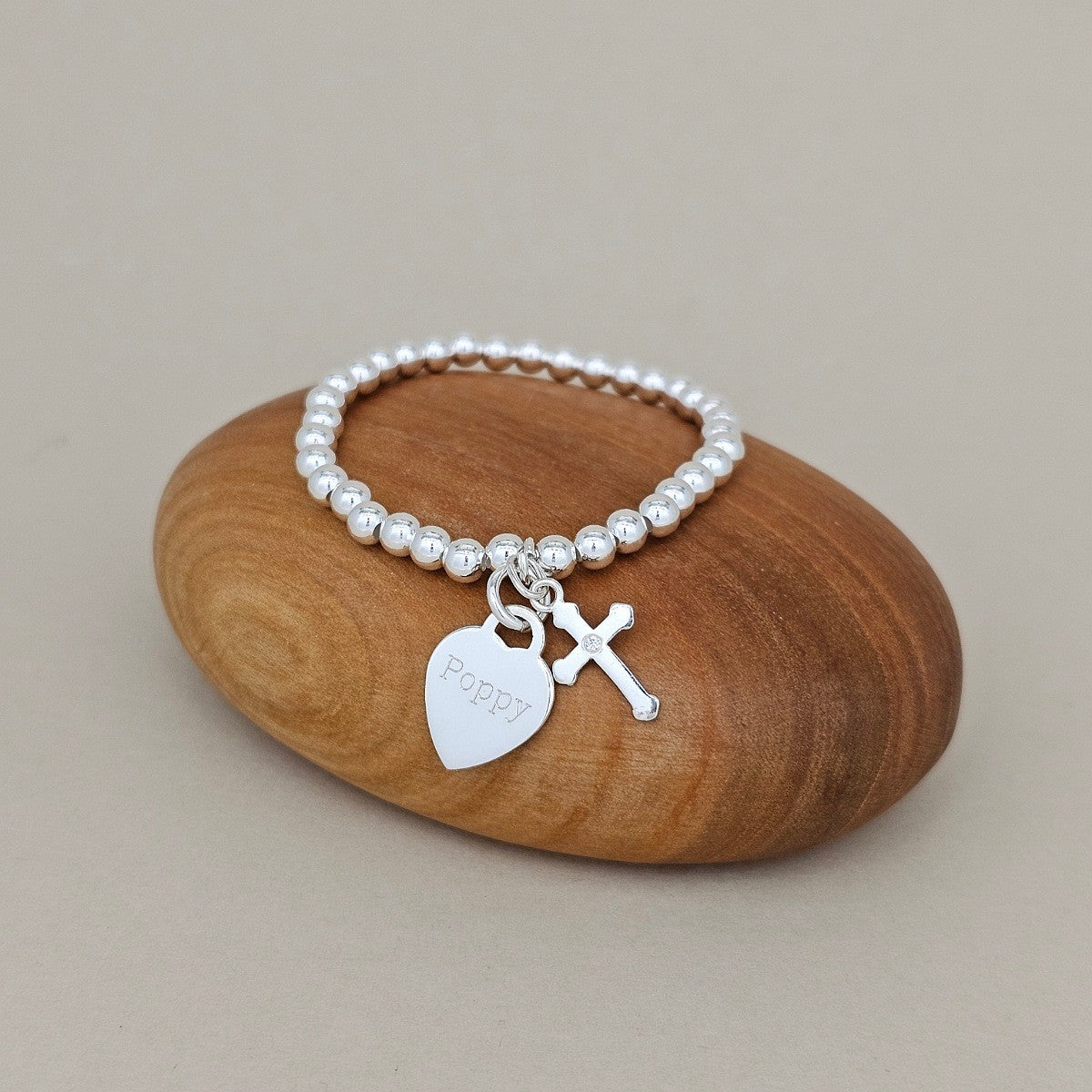 A silver beaded bracelet with a heart-shaped charm engraved with 'poppy' and a small cross charm, displayed on a smooth brown wooden stone against a neutral beige background.