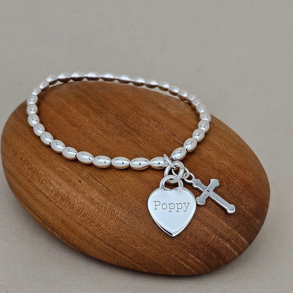 Silver bracelet with heart-shaped charm with the name 'Poppy' engraved and cross on a wooden rock
