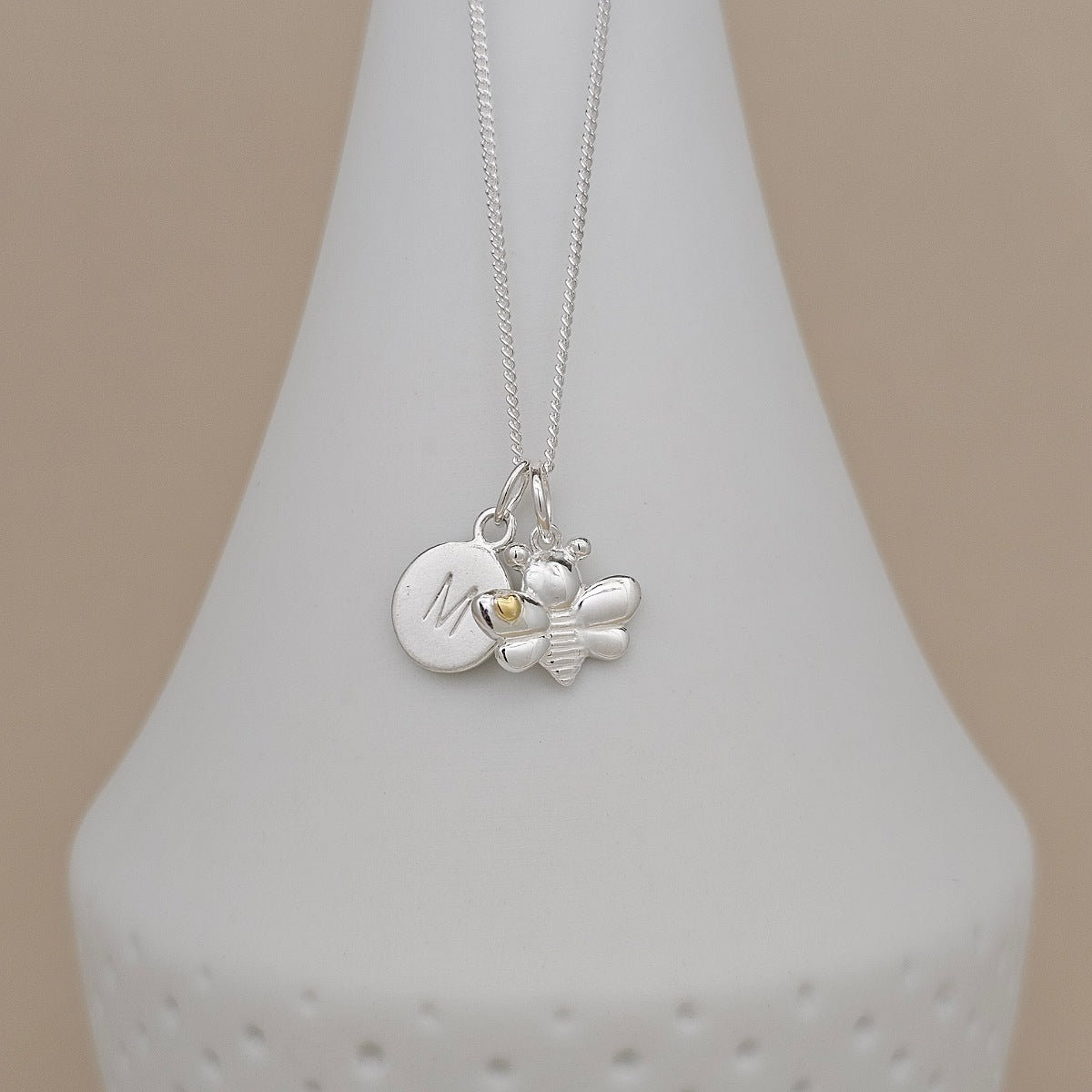A silver necklace with a round engraved initial 'M' disc charm and a small bee charm, displayed on a white cone-shaped stand against a beige background.