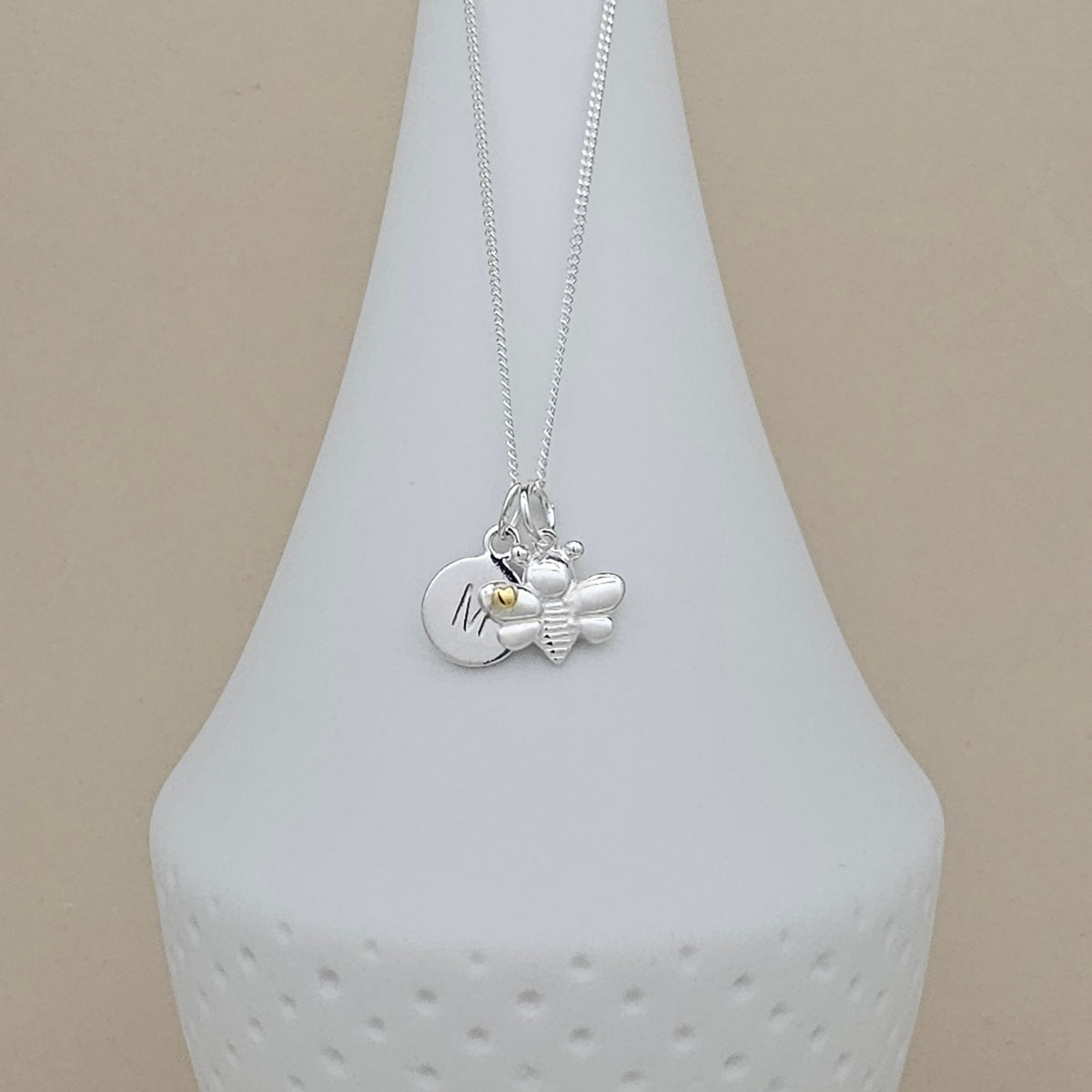 A silver pendant necklace featuring a round engraved disc and a small bee charm, displayed on a white ceramic vase against a beige background.