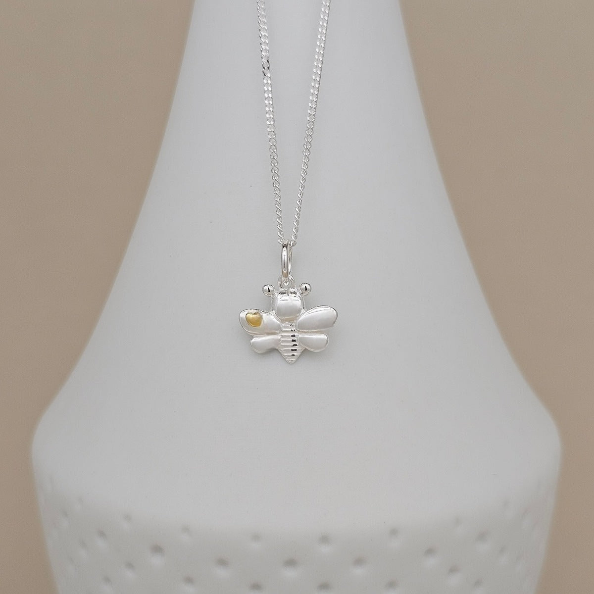 A silver bee pendant necklace with a small gold accent on the wing, displayed on a white ceramic jewelry bust against a beige background.