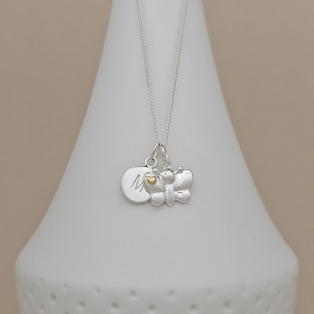 A silver pendant necklace displayed on a white cone-shaped jewelry stand against a warm beige background. The necklace features a silver butterfly charm, a round silver disc engraved with the letter 'M', and a tiny gold heart accent.