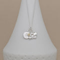 A silver pendant necklace displayed on a white cone-shaped jewelry stand against a warm beige background. The necklace features a silver butterfly charm, a round silver disc engraved with the letter 'M', and a tiny gold heart accent.