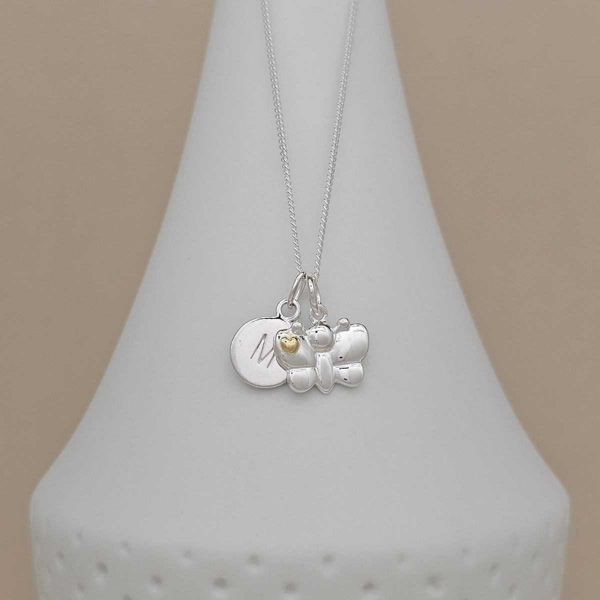 A silver pendant necklace displayed on a white cone-shaped jewelry stand against a warm beige background. The necklace features a silver butterfly charm, a round silver disc engraved with the letter 'M', and a tiny gold heart accent.