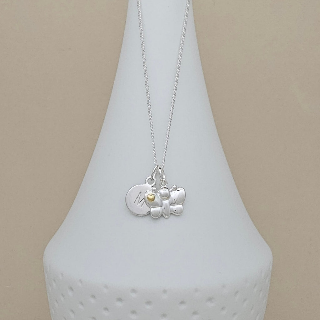 A silver pendant necklace displayed on a white ceramic vase against a beige background. The necklace features a silver chain and a multi-charm pendant including a round engraved disc, a butterfly charm, and a small yellow heart.