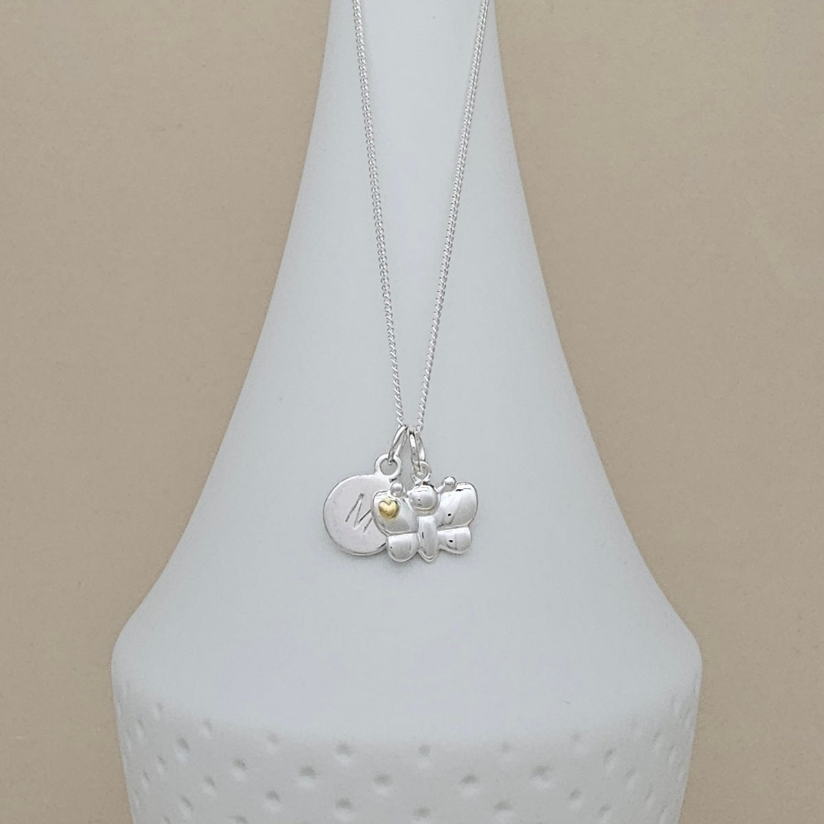 A silver pendant necklace displayed on a white ceramic vase against a beige background. The necklace features a silver chain and a multi-charm pendant including a round engraved disc, a butterfly charm, and a small yellow heart.