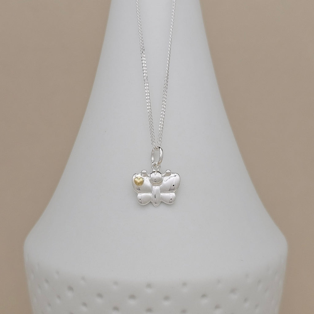 A silver butterfly pendant necklace with a small gold heart accent, displayed on a white ceramic vase against a soft beige background.
