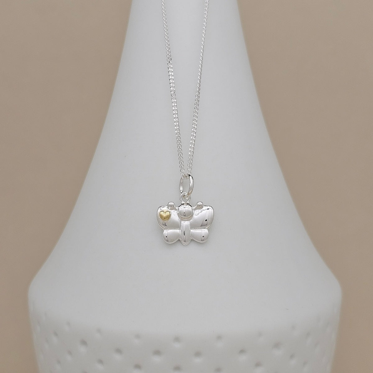 A silver butterfly pendant necklace with a small gold heart accent, displayed on a white ceramic vase against a soft beige background.