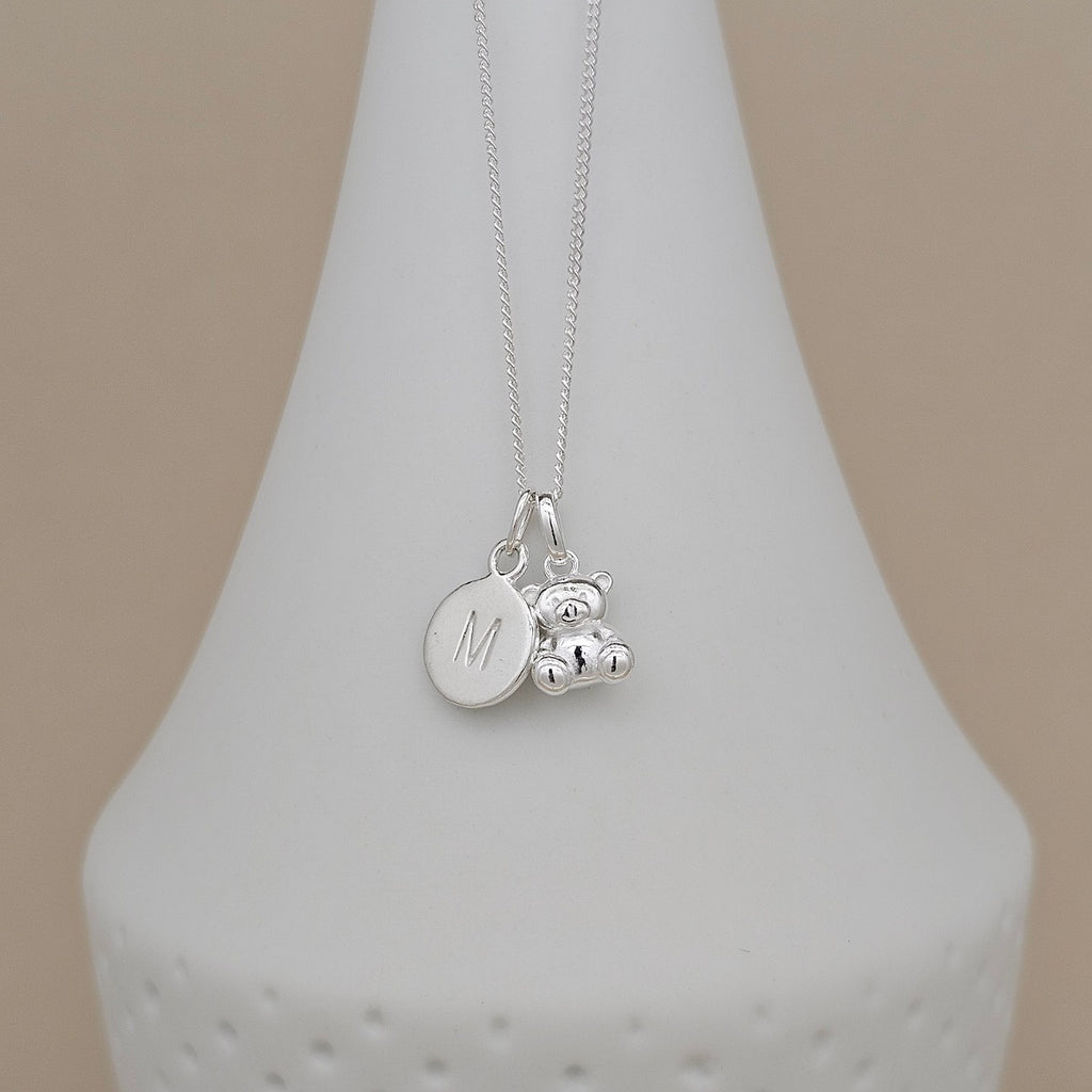 A silver necklace with a round engraved disc charm marked with the letter 'M' and a small teddy bear charm, displayed on a white cone stand against a beige background.