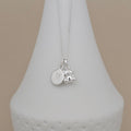 A silver necklace with a round engraved disc charm marked with the letter 'M' and a small teddy bear charm, displayed on a white cone stand against a beige background.