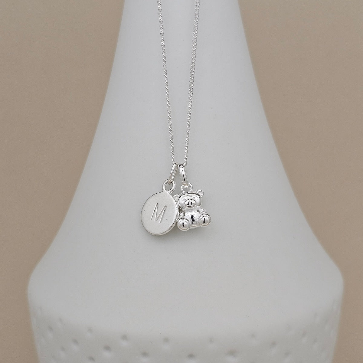 A silver necklace with a round engraved disc charm marked with the letter 'M' and a small teddy bear charm, displayed on a white cone stand against a beige background.