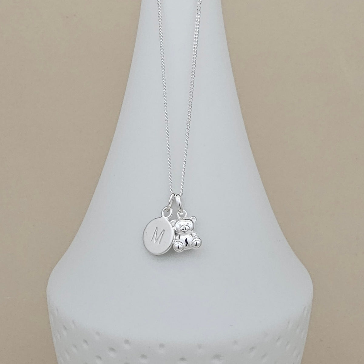 A silver necklace with a small teddy bear charm and a round engraved initial 'M' disc charm, displayed on a white ceramic vase against a beige background.