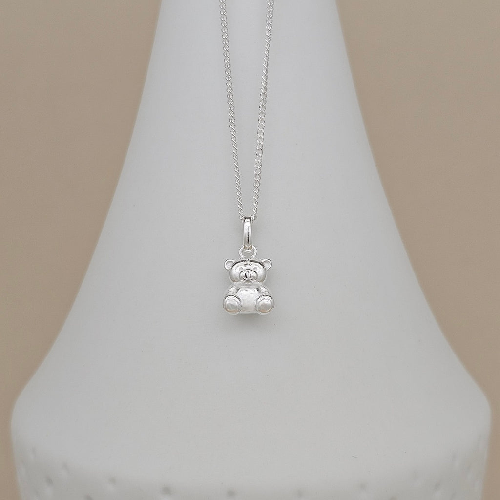 A silver teddy bear pendant necklace on a delicate silver chain, displayed on a white cone jewelry stand against a soft beige background.
