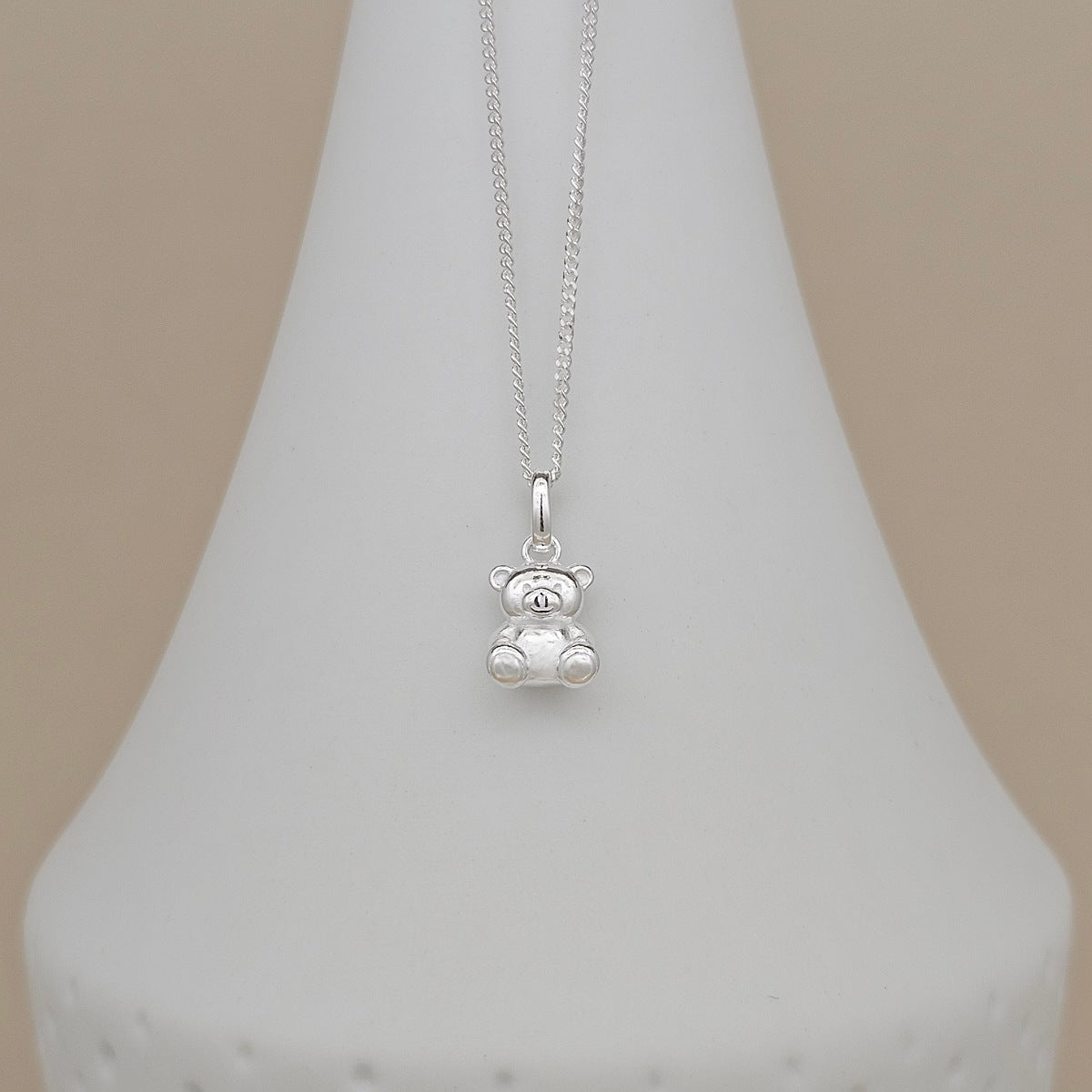 A silver teddy bear pendant necklace on a delicate silver chain, displayed on a white cone jewelry stand against a soft beige background.