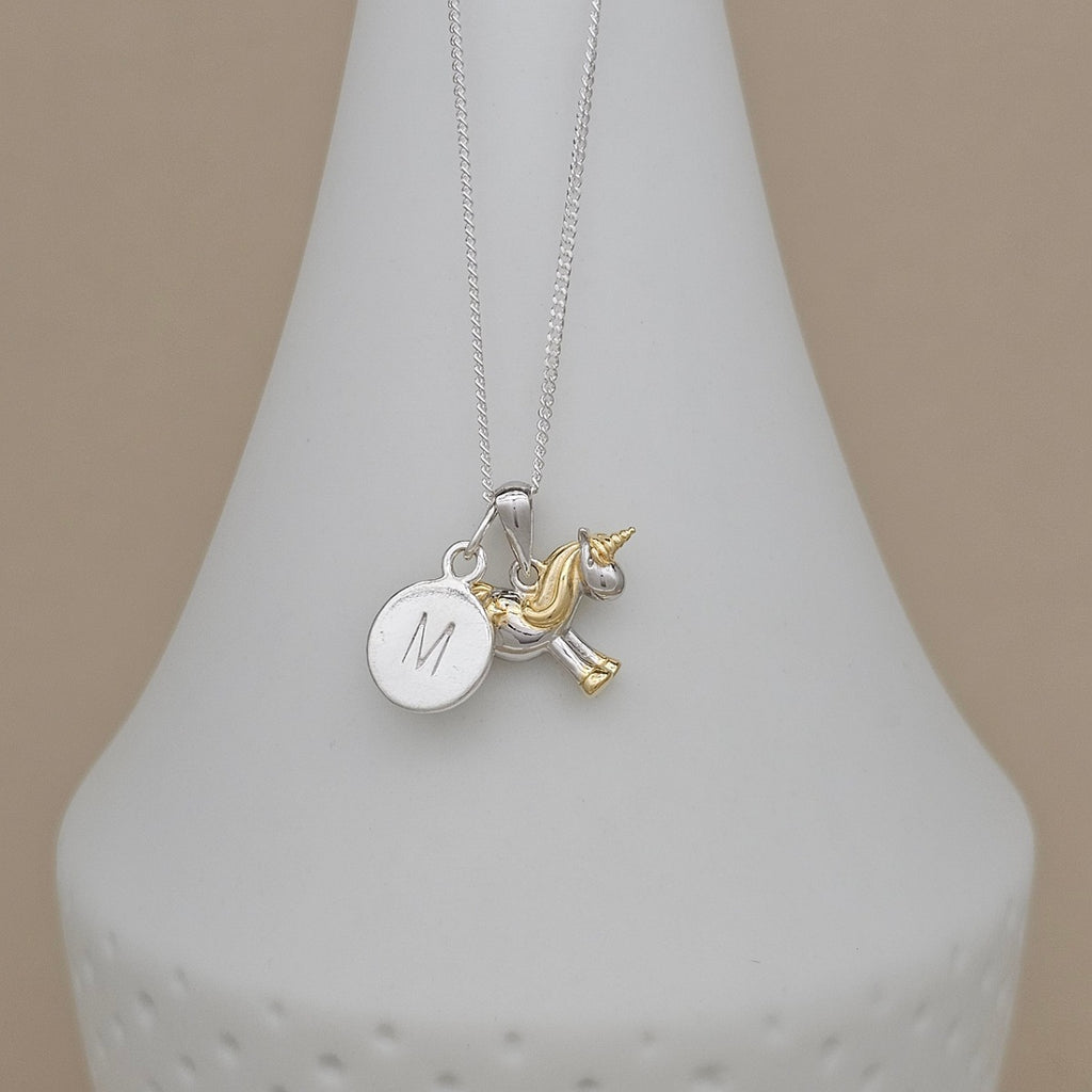 A silver necklace with a round engraved initial 'M' disc charm and a small gold unicorn charm, displayed on a white cone stand against a beige background.