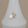 A silver necklace with a round engraved initial 'M' disc charm and a small gold unicorn charm, displayed on a white cone stand against a beige background.