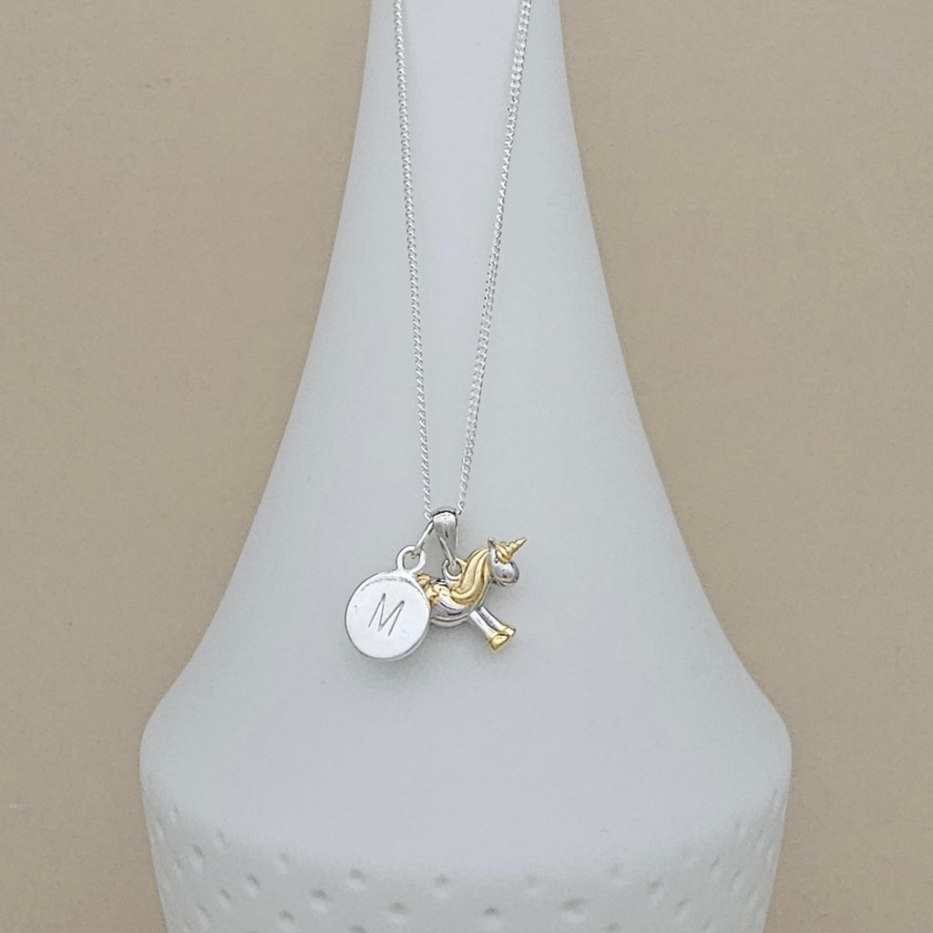 A silver chain necklace with a round engraved silver disc charm marked 'M' and a small gold unicorn charm, displayed on a white ceramic vase against a beige background.