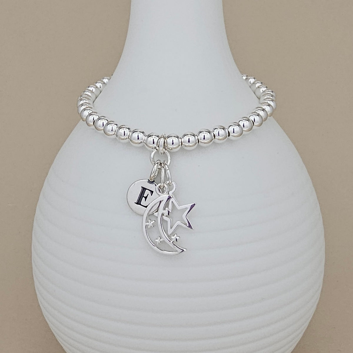A silver beaded bracelet with a crescent moon, star, and initial 'E' charm, displayed on a white ribbed ceramic vase against a beige background.