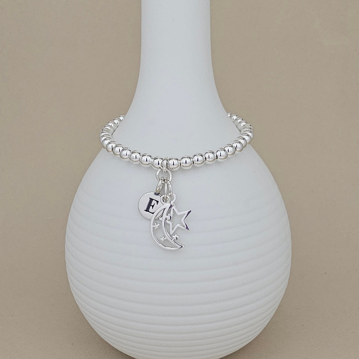 A silver beaded bracelet with a crescent moon, star, and initial 'E' charm, displayed on a ribbed white ceramic vase against a beige background.