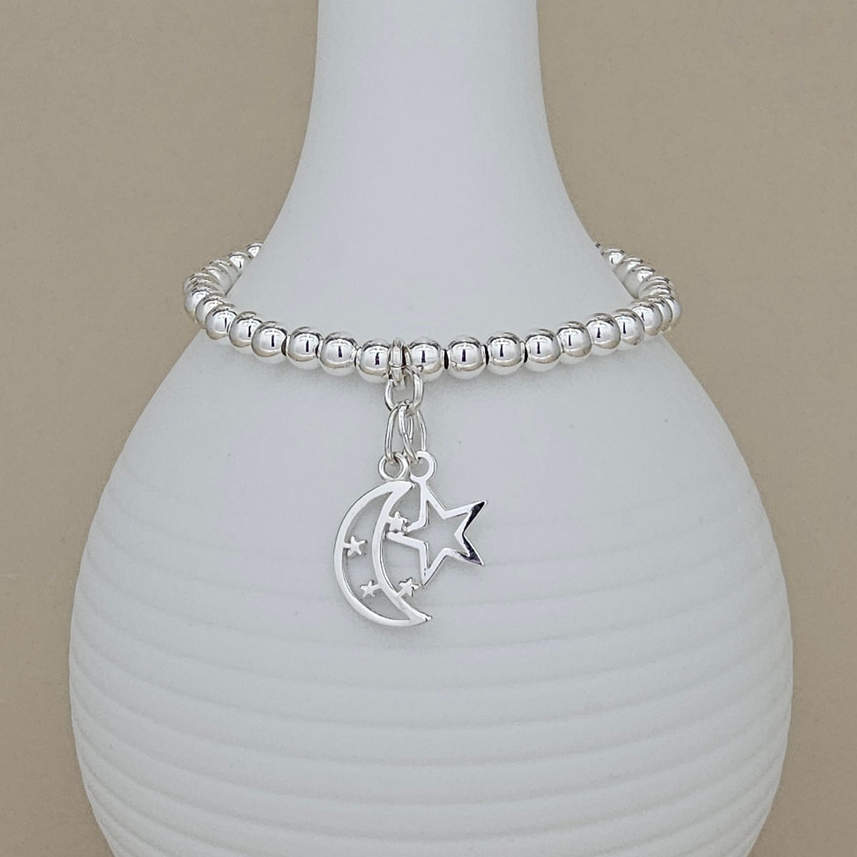 A silver beaded bracelet with a crescent moon and star charm, displayed on a white ribbed ceramic vase against a beige background.