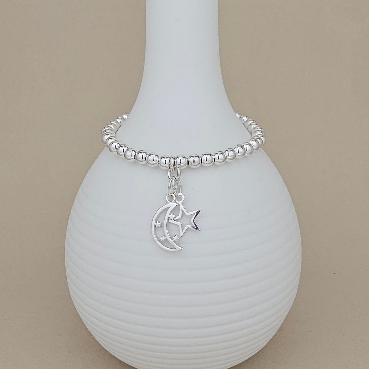 A silver beaded bracelet with a crescent moon and star charm, displayed on a ribbed white ceramic vase against a beige background.